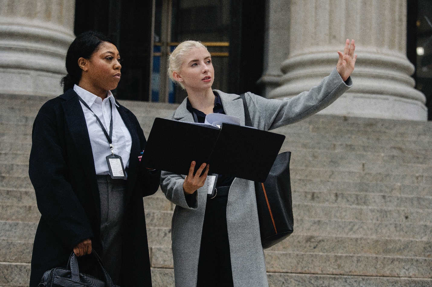 Two women stand on stairs leading up to a public building, discussing something while looking at a file