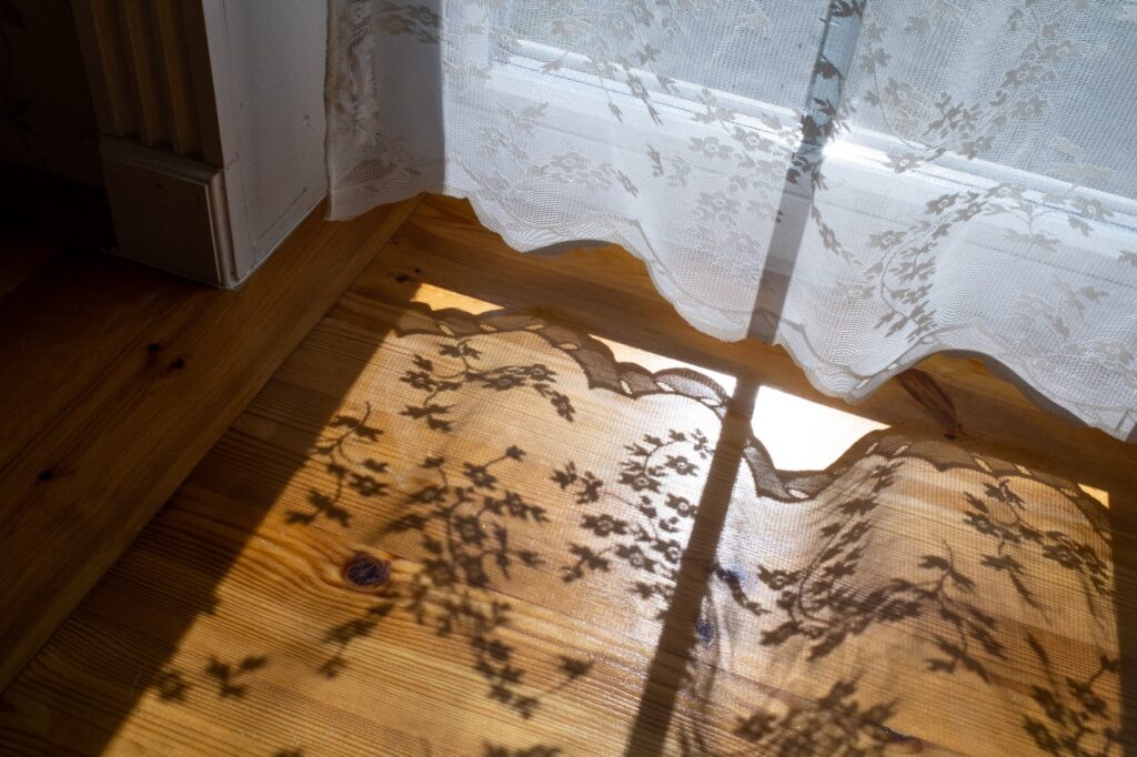 The shadow of a white net curtain with flowers falls upon a hardwood floor