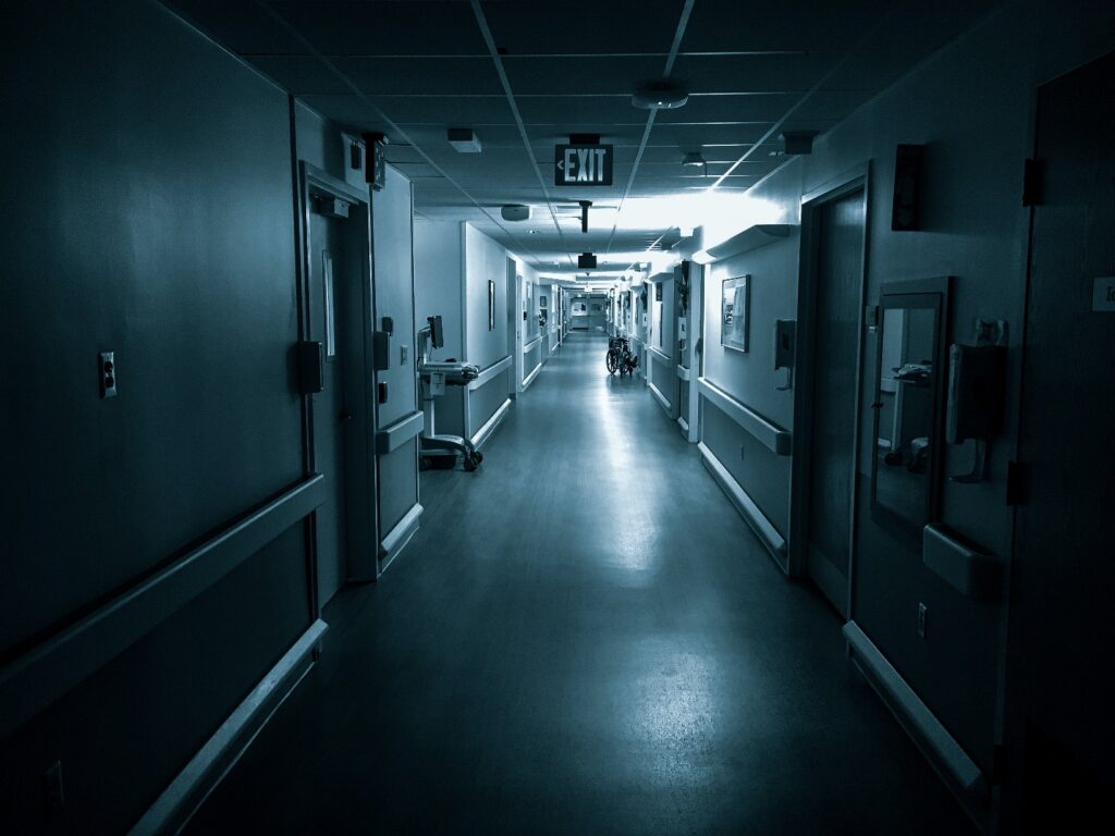 A dark hospital hallway with doors on either side and a patient in a wheelchair further down