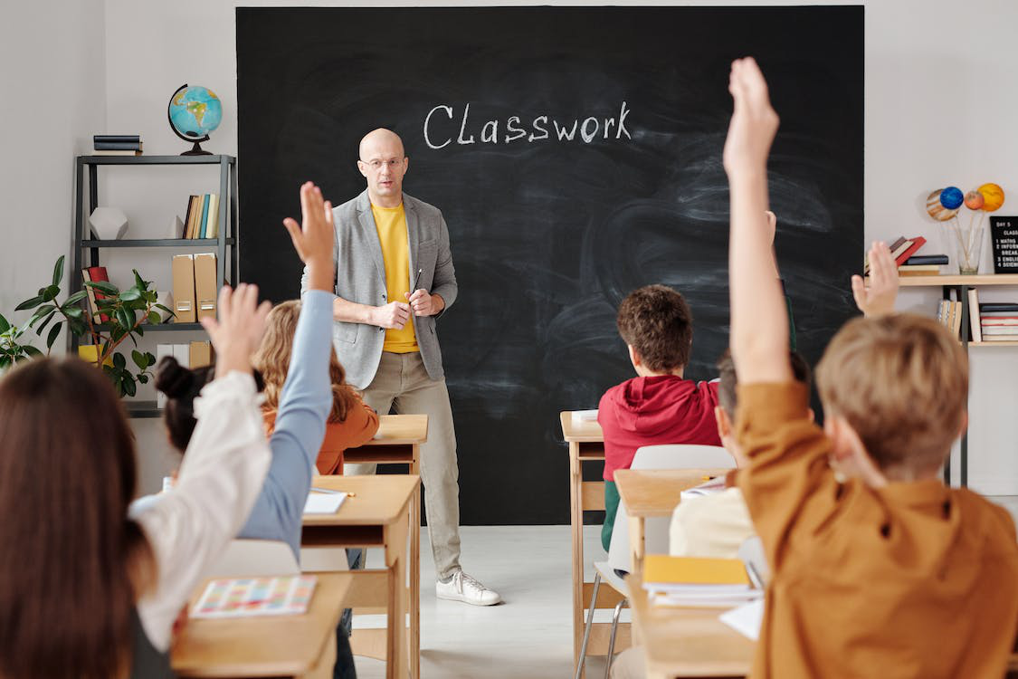 Students raising their hands to participate in class discussion while the teacher stands near the board