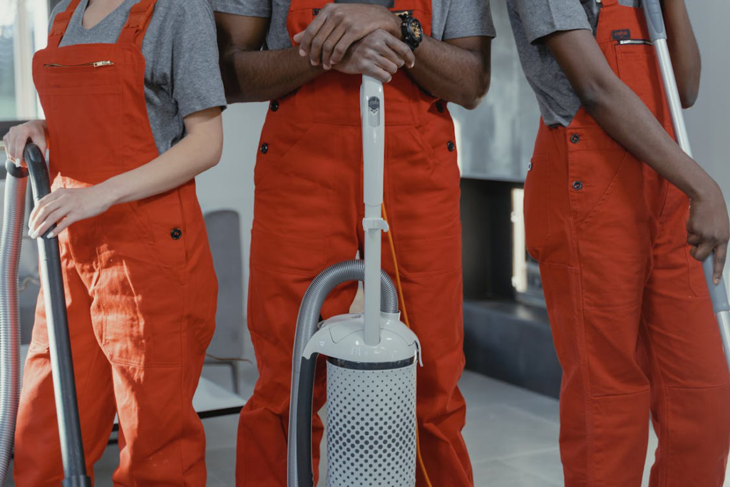 An image of three janitors in orange overalls holding cleaning equipment