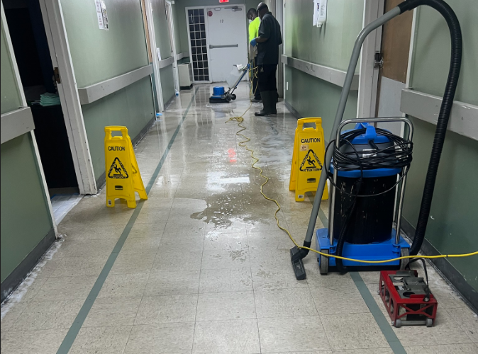 Two workers are cleaning a corridor with caution signs and cleaning equipment placed along the floor.